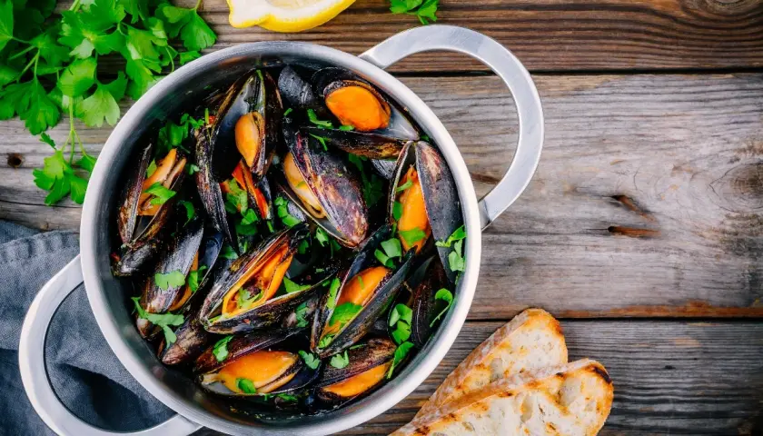 Un pentolino pieno di cozze alla marinara, guarnite con prezzemolo fresco e accompagnate da fette di pane tostato e spicchi di limone.
