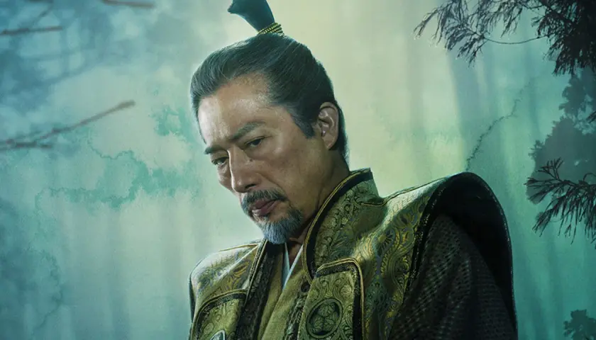 En äldre man med gråsprängt skägg och en tofs i håret, klädd i en utsmyckad dräkt. En karaktär från shogun serie.