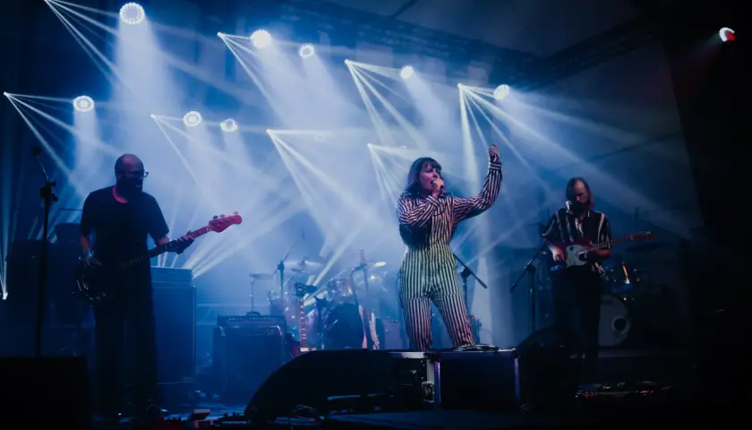 Izzy i The Black Trees na scenie, otoczeni niebieskimi światłami. Wokalistka w pasiastym stroju śpiewa, gitarzyści grają.