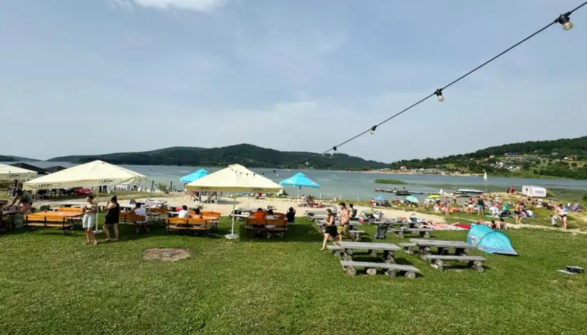 Słoneczny dzień nad Jeziorem Mucharskim. Ludzie odpoczywają na plaży, pod parasolami, przy stolikach. W tle jezioro i wzgórza. Atrakcje dla każdego.