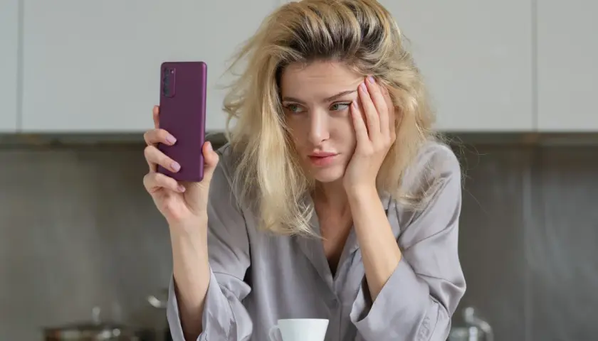 Zmęczona kobieta z telefonem w dłoni, z ręką na twarzy. Czy to objawy uzależnienia od kofeiny?