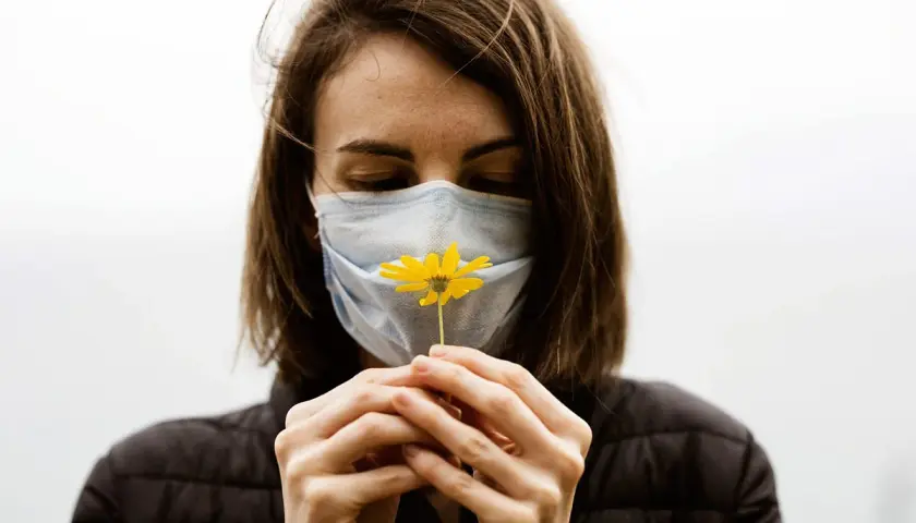 Mujer con mascarilla azul, oliendo una flor amarilla. Diversos tipos de mascarillas protegen, pero la esperanza florece.
