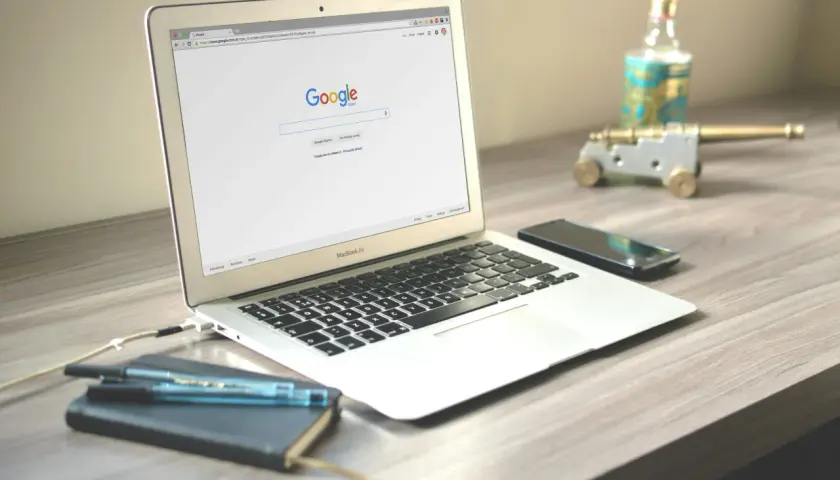 Laptop z otwartą stroną wyszukiwarki Google. Obok leży smartfon, notatnik z długopisami i ozdobne działko.