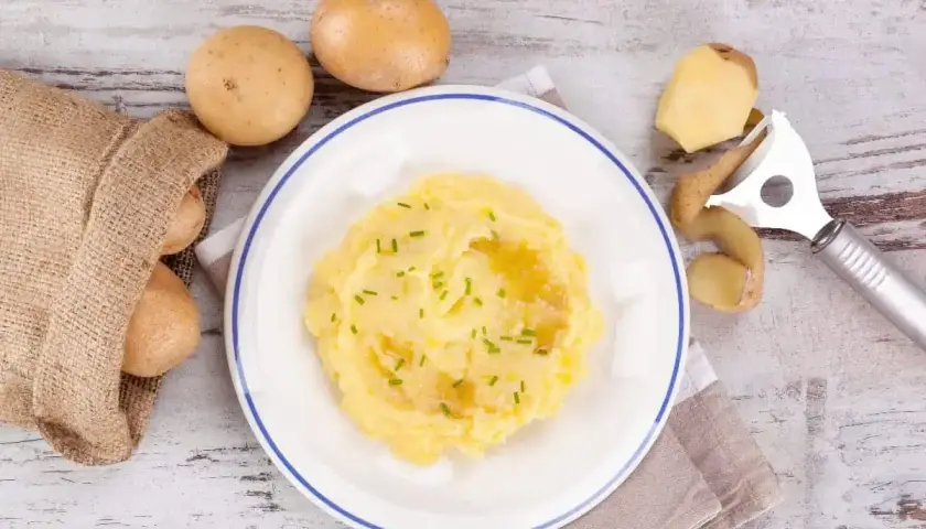 Pyszne puree ziemniaczane na talerzu, obok surowe ziemniaki i obieraczka. Czy cukrzyk może jeść ziemniaki? Tak, w umiarkowanych ilościach!