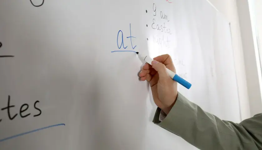 Mano scrive "at" su lavagna, spiegando il suo significato in inglese.