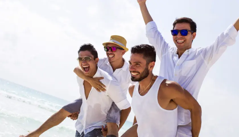 Czterech roześmianych mężczyzn na plaży, jeden niesie drugiego na barana. Wyglądają na to, kto organizuje wieczór kawalerski, świętując na wakacjach.