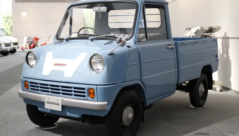 Niebieski, zabytkowy pick-up Honda z dużym białym "H" na masce. Ten model, choć nie jest Isuzu, przypomina klasyczne japońskie pojazdy użytkowe.
