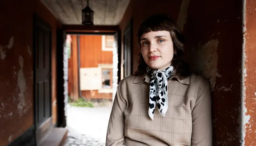 Sanna Samuelsson står i en gång med orange väggar och en träfasad i bakgrunden. Hon bär en beige tröja och en scarf med koskinnmönster.