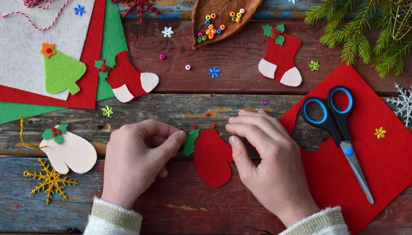 Manos creando adornos navideños con fieltro rojo y verde. Tijeras, cuentas y piñas completan la escena de manualidades.