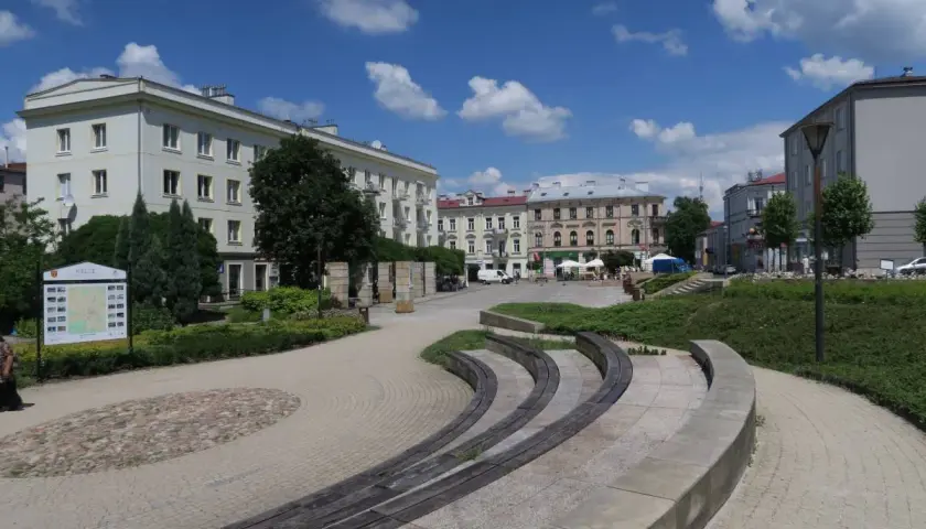 Słoneczny dzień na placu artystów w Kielcach. Widać budynki, zieleń i ławki.