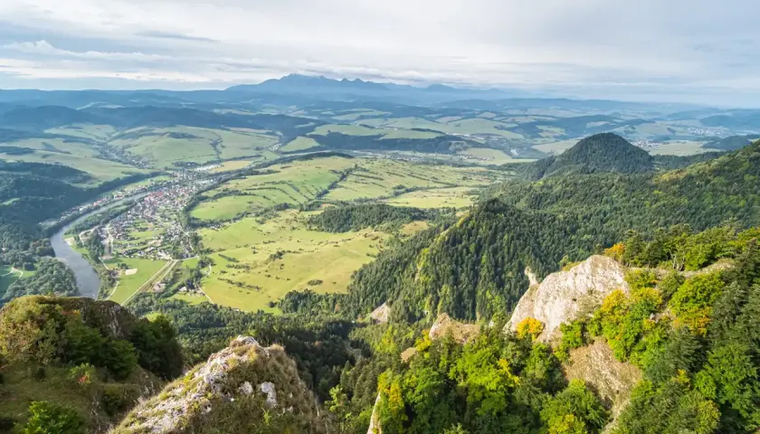 Widok z góry na dolinę z rzeką i miasteczkiem, w oddali majaczą Tatry. Widać skaliste zbocza porośnięte lasem, a w tle trzy korony nazwy szczytów.