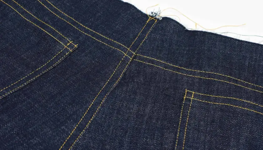 Close-up of the yoke on dark blue jeans, showing yellow stitching and raw edges.
