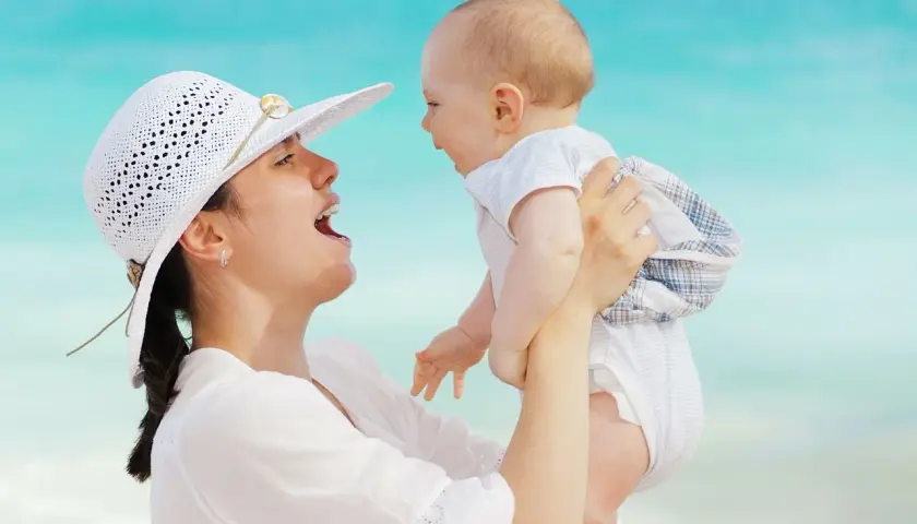 Mamma solleva il suo neonato di 5 mesi, forse le prime parole sono in arrivo, con il mare azzurro sullo sfondo.