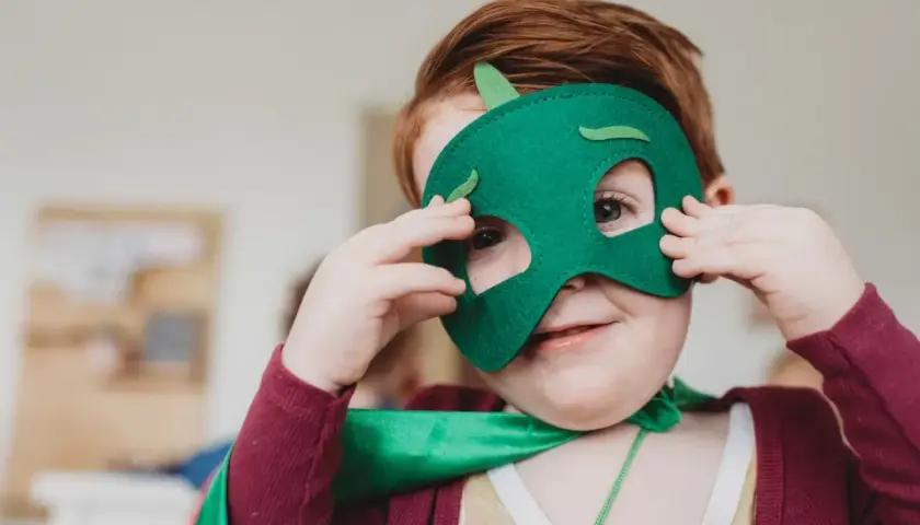 Ein Kind mit roten Haaren trägt eine grüne Drachenmaske und einen Umhang. Das Kind schaut neugierig durch die Augenlöcher.