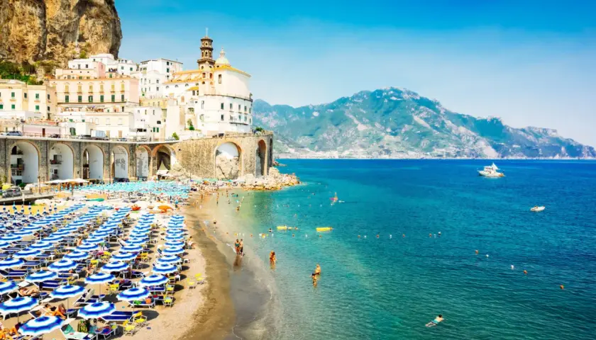 A vibrant beach scene in Naples, with rows of blue umbrellas and sunbathers. Buildings cling to a cliffside, overlooking the sparkling turquoise sea.