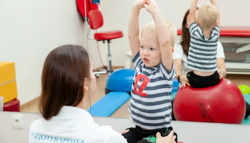 Fizjoterapeutka wspiera chłopca z zaburzeniami rozwojowymi podczas ćwiczeń na piłce rehabilitacyjnej.