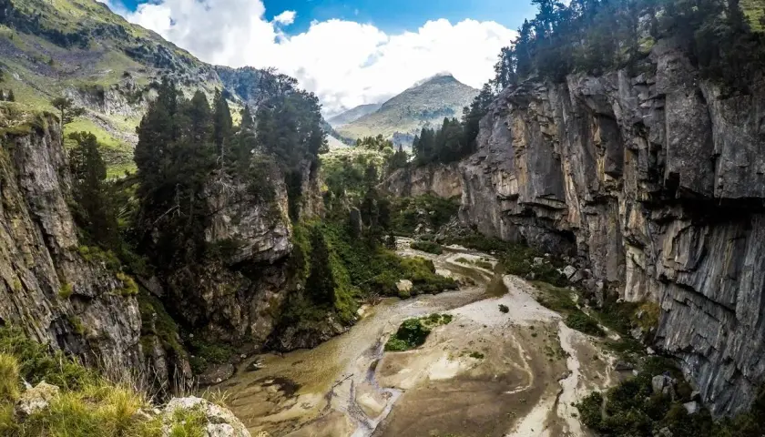 El forau de Aigualluts desde los Llanos del Hospital, un cañón rocoso con un río seco y vegetación verde bajo un cielo azul con nubes.