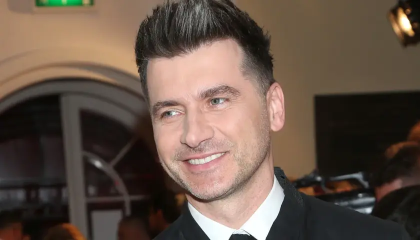 Uśmiechnięty Tomasz Kammel, jego wiek nie jest widoczny, ale jego stylizacja i fryzura sugerują, że jest w sile wieku.