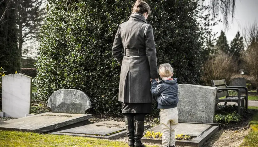 Kobieta z dzieckiem na cmentarzu. Dziecko trzyma matkę za rękę, wspólnie przeżywają żałobę po babci.