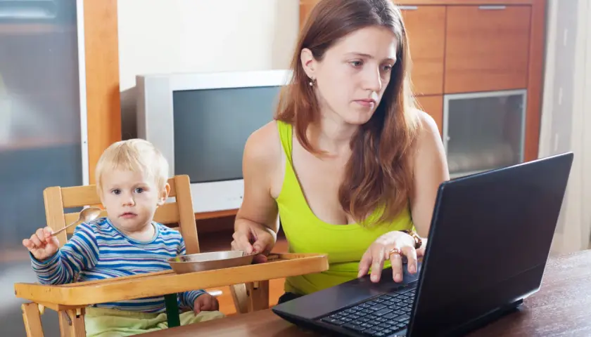 Mama na zasiłku macierzyńskim pracuje przy laptopie, podczas gdy jej dziecko siedzi w krzesełku i trzyma łyżeczkę.