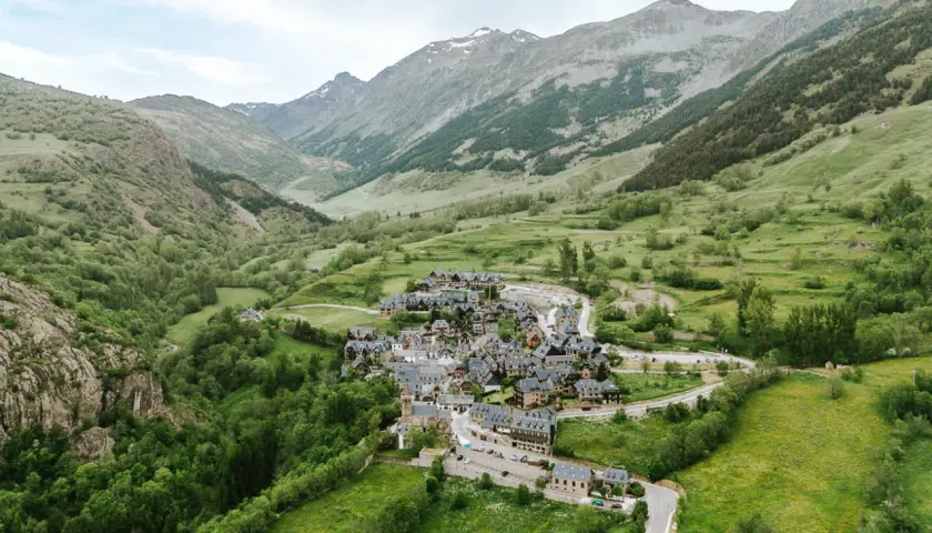 Pueblo de casas de piedra en un valle verde del Val d'Aran, rodeado de montañas cubiertas de vegetación y algunas cumbres nevadas.