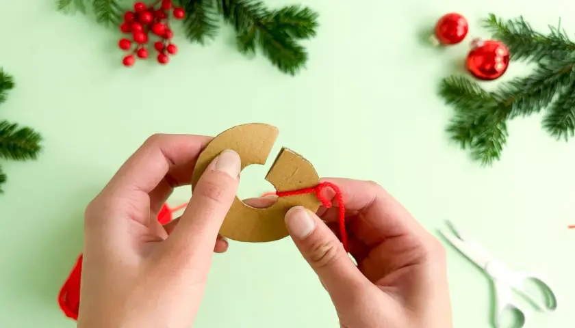 Hände wickeln roten Faden um Pappring für **Basteln mit Pompons Weihnachten**. Tannenzweige und Kugeln im Hintergrund.