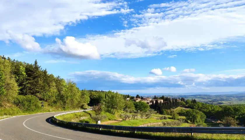 Strada SR 222 Chiantigiana serpeggia tra colline verdi e boschi, con un borgo toscano in lontananza sotto un cielo azzurro e nuvoloso.