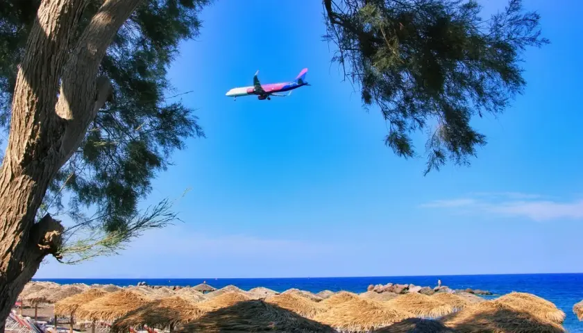 Samolot przelatuje nad plażą z parasolami, tuż przed lądowaniem na **Santorini lotnisko**.