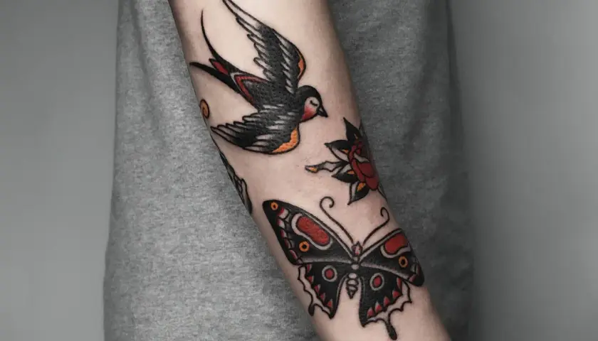 Kolorowy tatuaż tradycyjny na przedramieniu: lecąca jaskółka, motyl i róża.