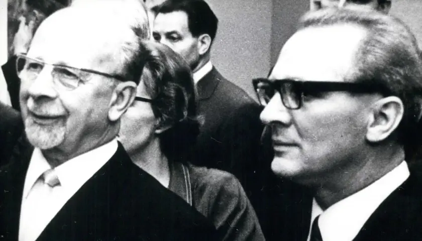 Ein Mann mit Brille und Bart, ein DDR-Chef, lächelt. Im Hintergrund weitere Personen.