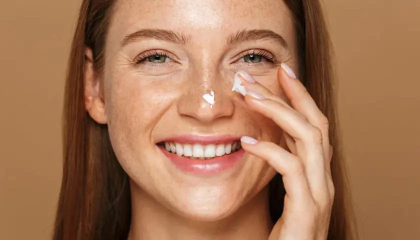 Uśmiechnięta kobieta z piegami aplikuje krem na nos. Pamiętaj o ochronie przed słońcem, zwłaszcza stosując retinol.