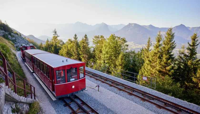 Czerwony wagon kolejki zębatej jedzie po torach w górę zbocza. W tle góry i lasy.