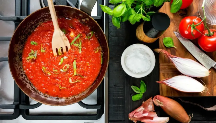 En kastrull med bubblande tomatsås och en träsked. Ingredienser för ett tomatsås recept: färska tomater, schalottenlök, basilika och salt.