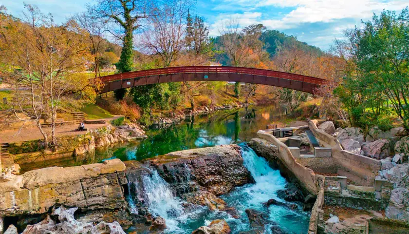 Cascada y puente de madera en la vía verde Puente Viesgo, rodeados de árboles otoñales y un cielo azul vibrante.