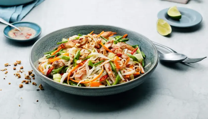 En läcker asiatisk nudelsallad med kyckling, grönsaker och jordnötter. Serveras med en dressing och limeklyftor.