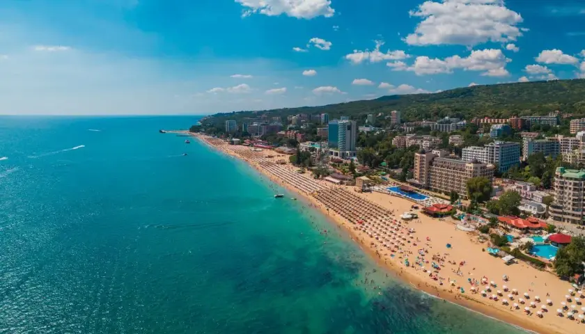 Słoneczna plaża z mnóstwem leżaków i parasoli, tętniące życiem kurorty nad morzem, a w oddali zielone wzgórza. Idealne miejsce na wakacje.