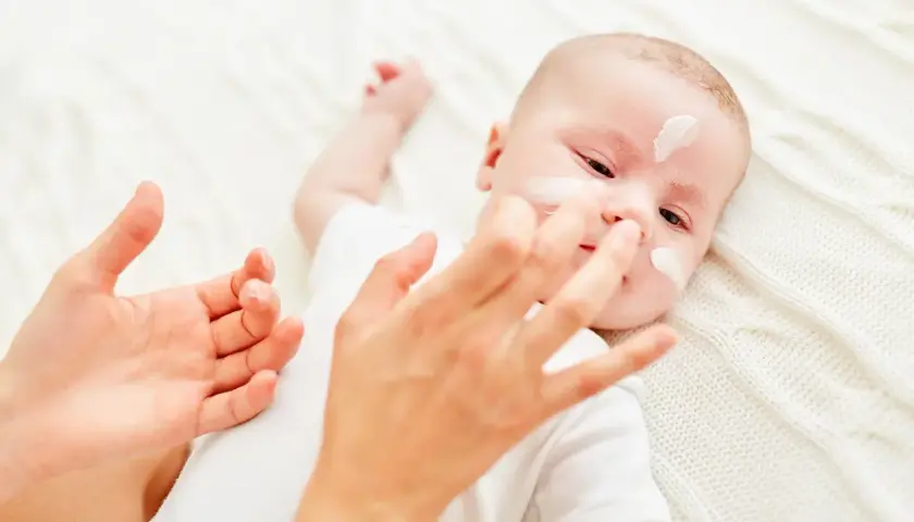 Delikatna pielęgnacja noworodka. Krem na buzi maluszka, troskliwe dłonie mamy.
