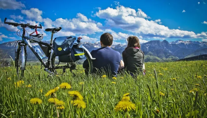 Para na rowerach odpoczywa na łące pełnej mleczy, podziwiając Tatry. To idealne miejsce na szlak papieski Gubałówka.