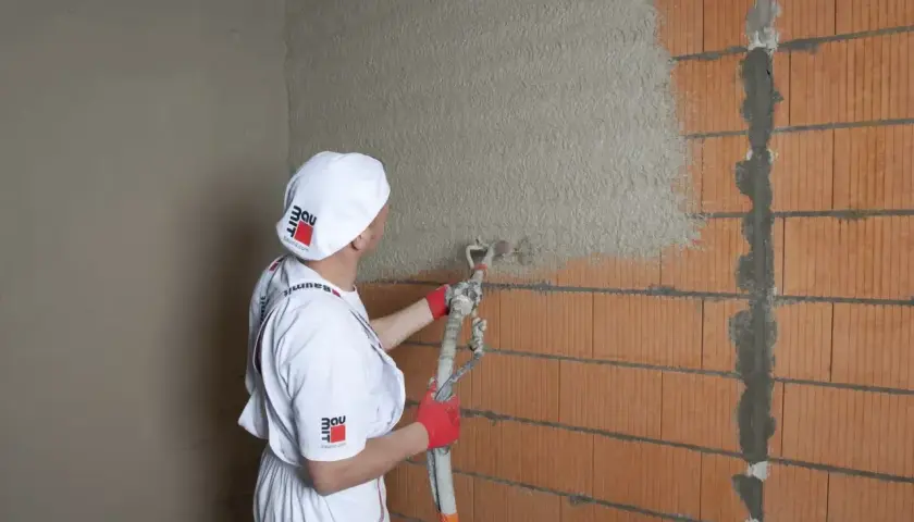 Pracownik w białym stroju nakłada tynki cementowo wapienne na ścianę z pustaków ceramicznych za pomocą agregatu.
