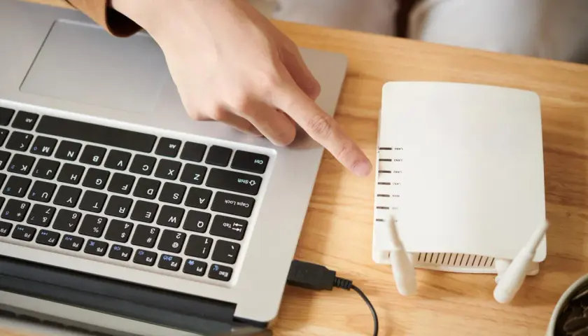 Dłoń wskazuje na router, podłączony kablem do laptopa. Dowiedz się, jak włączyć wifi w laptopie, korzystając z tego połączenia.