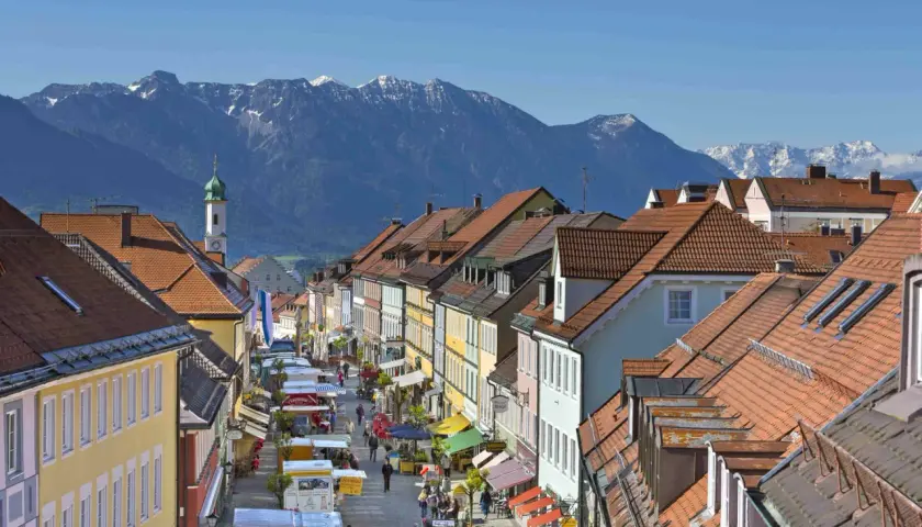 Bunte Häuserfronten säumen eine belebte Straße mit Marktständen, im Hintergrund majestätische Berge. Entdecke Murnau Geheimtipps!