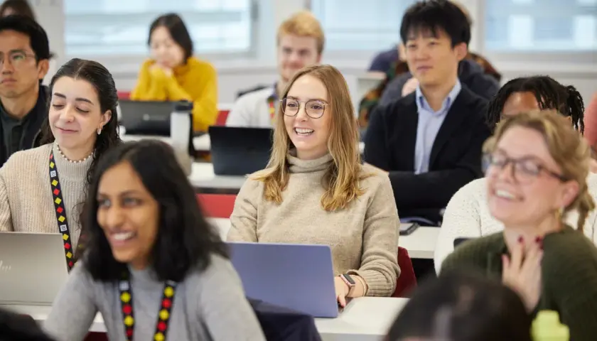Students in a classroom, some with laptops, are engaged in a lesson. One student smiles, perhaps anticipating her MPA credential.