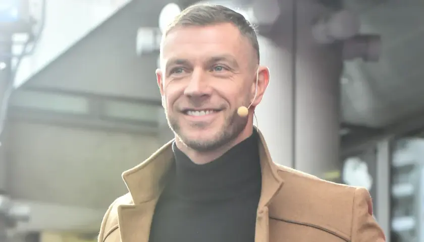 Uśmiechnięty Dariusz Pachut w beżowym płaszczu i czarnym golfie, z mikrofonem przy uchu. Może opowiada o swojej żonie.
