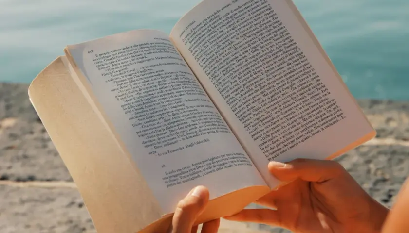 Mani che sfogliano un libro aperto, con un mare azzurro sullo sfondo. Un'immagine che evoca la lettura di una **canzone in dialetto siciliano** sotto il sole.