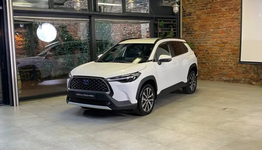 Biała Toyota Corolla Cross Hybrid stoi w nowoczesnym wnętrzu z ceglaną ścianą i dużymi oknami.