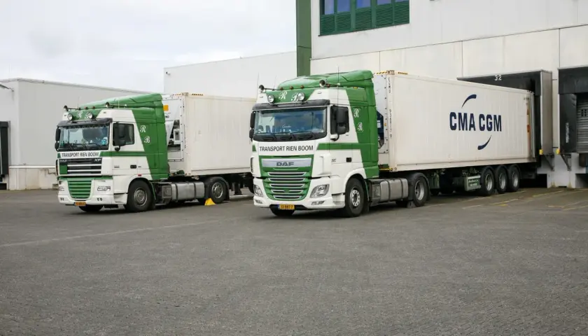 Zwei weiße und grüne LKWs stehen bereit, um entladen zu werden. Einer trägt die Aufschrift "Transport Rien Boom", der andere "CMA CGM".