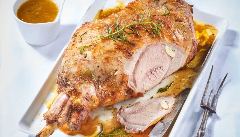 Gigot d'agneau désossé au four, doré et juteux, garni de romarin et d'ail. Servi avec sa sauce dans un plat blanc.