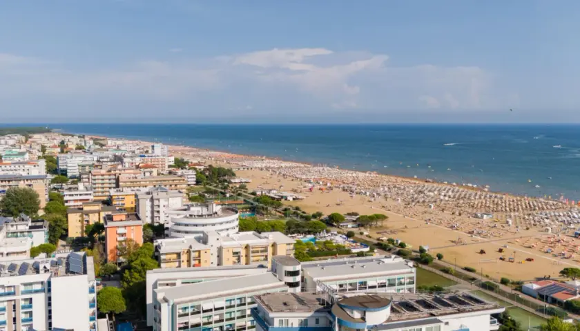 Widok z lotu ptaka na zatłoczoną bibione plażę, pełną białych parasoli i ludzi. W tle błękitne morze i nadmorskie miasteczko.