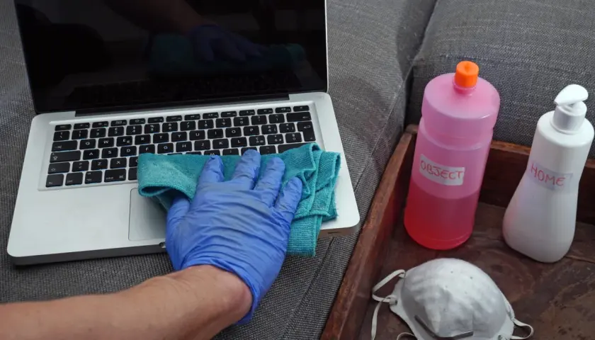 Ręka w niebieskiej rękawiczce czyści laptopa. Obok butelka z napisem "OBJECT" i maseczka. Denaturat jest do dezynfekcji przedmiotów.
