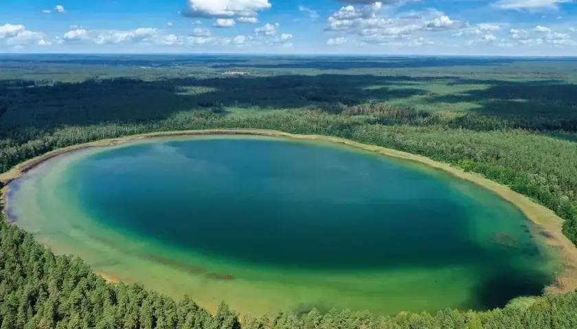 Widok z lotu ptaka na **jezioro płaskie** otoczone lasem. Woda ma turkusowy odcień, a brzegi są porośnięte drzewami.